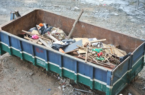 A large, rectangular metal skip positioned on a rough, gravel surface, filled with a variety ofConstruction debris and waste materials including wooden planks, broken pieces of wood, plastic bags, and miscellaneous rubbish. The skip has a dark rusted interior and a blue exterior with a green stripe running along the middle, indicating it is used for construction or garden clearance. Surrounding the skip is an outdoor area with uneven ground and scattered dirt, suggesting an active site for rubbish removal or renovation work. The environment appears to be a construction or garden clearance site, typical of what Garden Clearance Merton may serve, with the skip ready for waste disposal. The overall scene emphasizes waste collection and outdoor rubbish removal, supporting professional services for clearing garden or building debris in the Merton area or nearby locations.