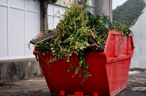 A large red plastic skip filled with garden waste, including branches, leafy green plants, and small twigs, situated on a paved outdoor area with a white fence and a concrete wall in the background. The garden debris is piled high and appears freshly collected, with some green foliage and white flowering plants visible among the cut branches. The skip is positioned on the driveway or patio surface, which is textured concrete. The setting suggests a garden clearance task, typical of outdoor rubbish removal services provided by Garden Clearance Merton in the local area. The lighting is natural, casting minimal shadows, and the scene captures the typical scene of garden refuse ready for disposal or recycling, emphasizing the type of waste managed during outdoor waste removal in Merton.